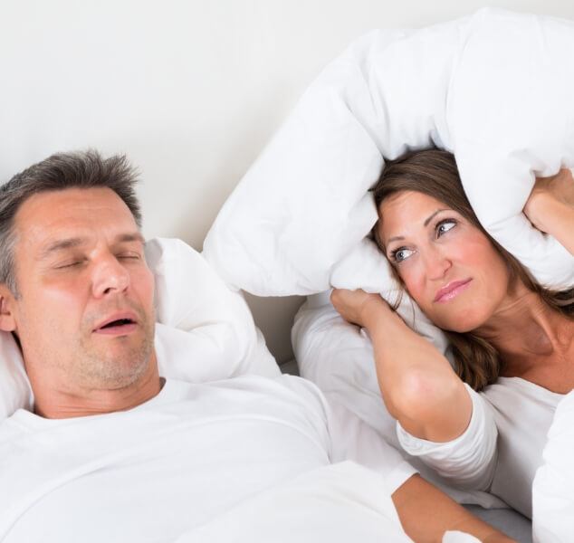 Woman in bed covering her ears with pillow next to sleeping man